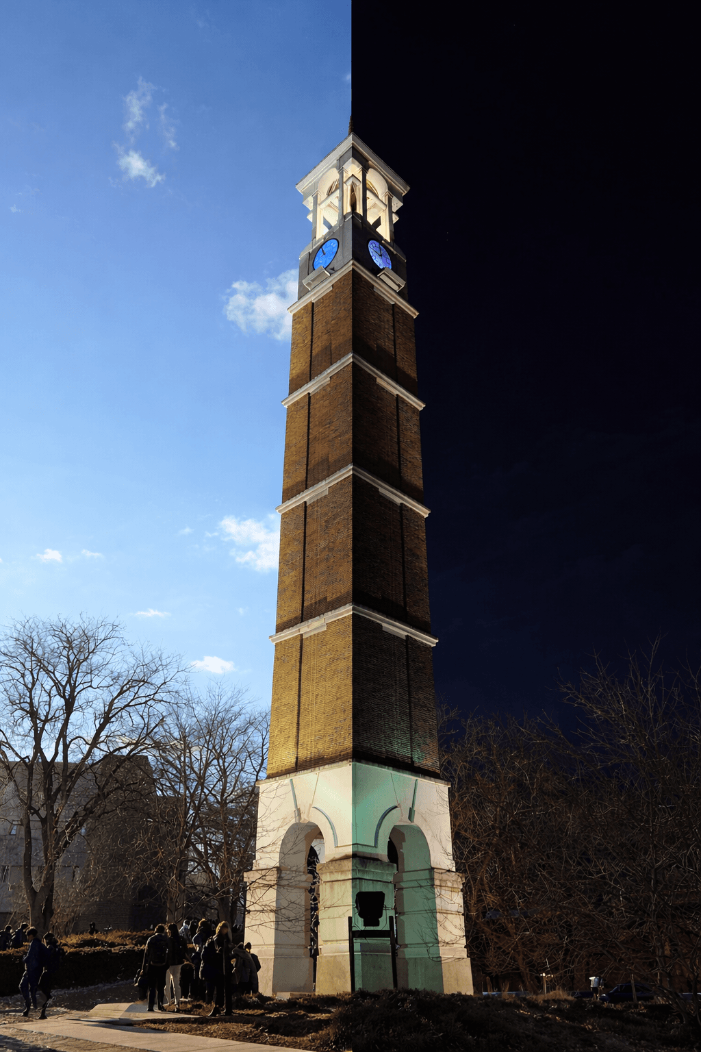 Split vertical composite of the same tall brick clock tower—left half in daylight with blue sky and students near the base, right half at night with blue-lit clock faces and green spotlit stone arches