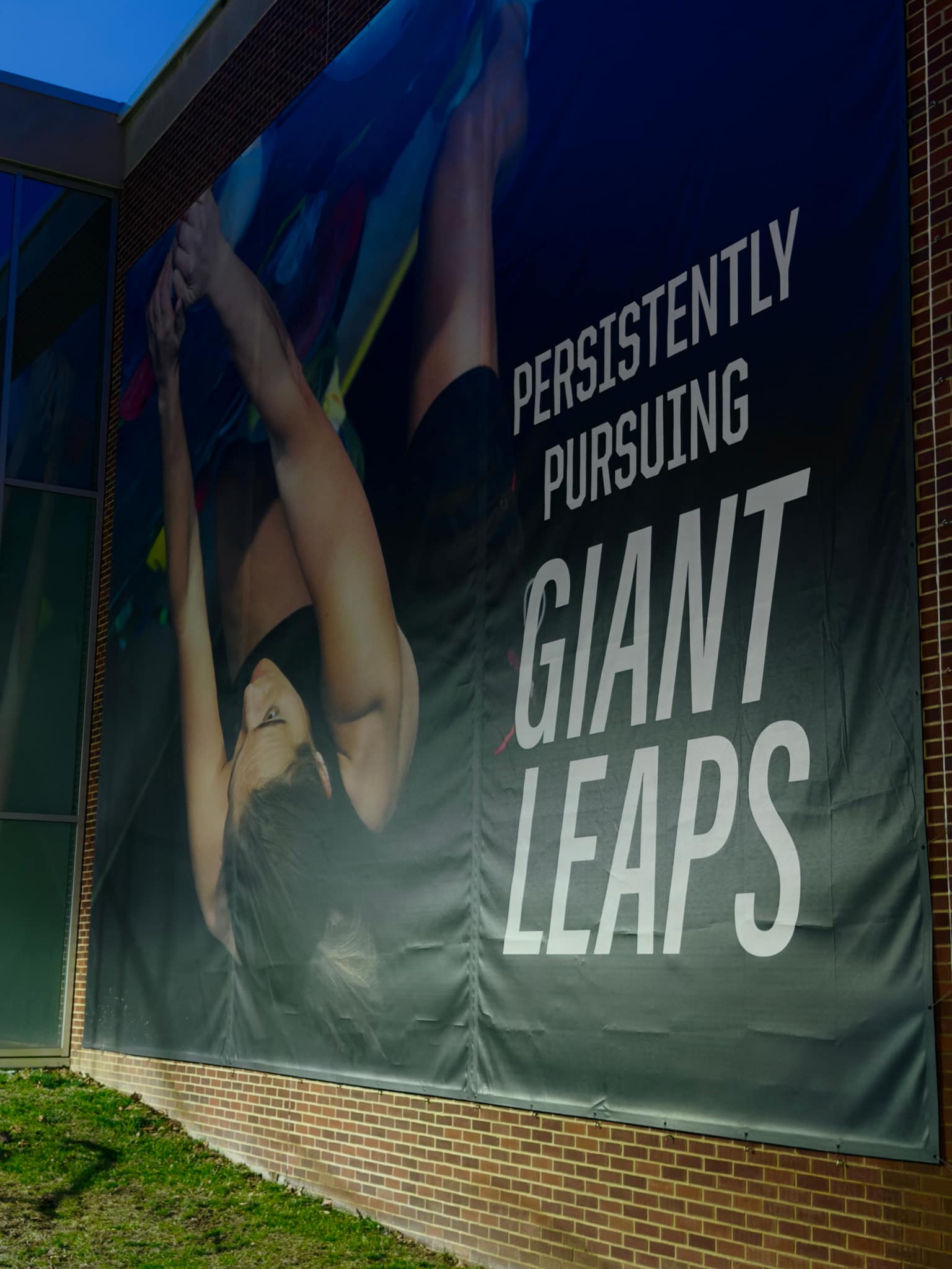 Tall building banner on red brick: navy field with white words Persistently Pursuing Giant Leaps beside a photo of a gymnast mid-motion, seen from a low angle with glass windows above