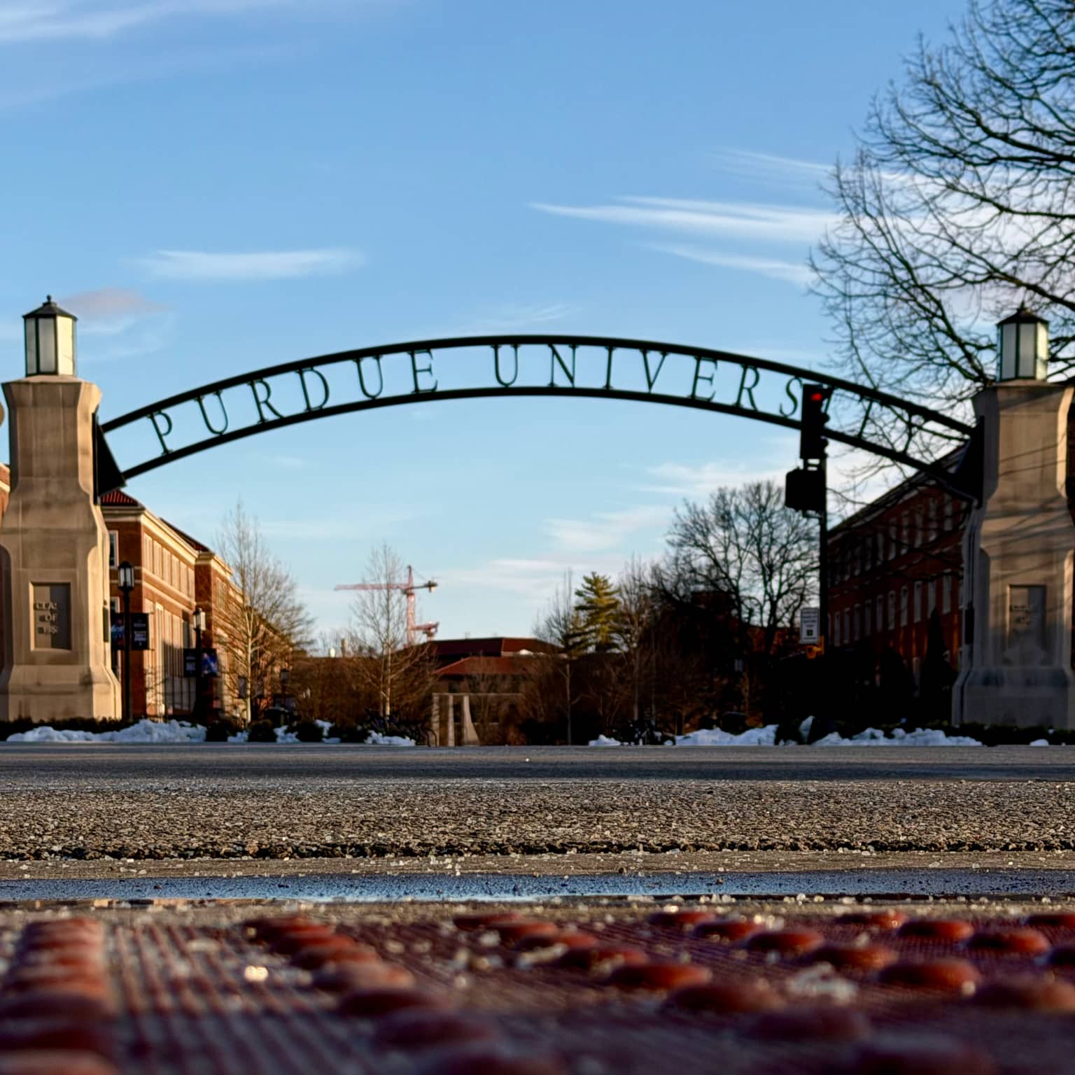 Low-angle view from the road looking up at the Purdue University gateway arch with white serif lettering, stone pillars and lanterns, bare trees and snow patches under a bright winter sky