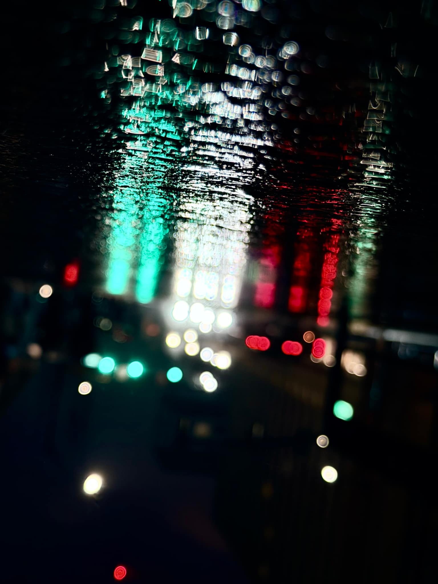 Abstract night photograph of vertical green, white, and red light columns blurred into soft waves with scattered multicolor bokeh, like reflections on wet glass or rippled water