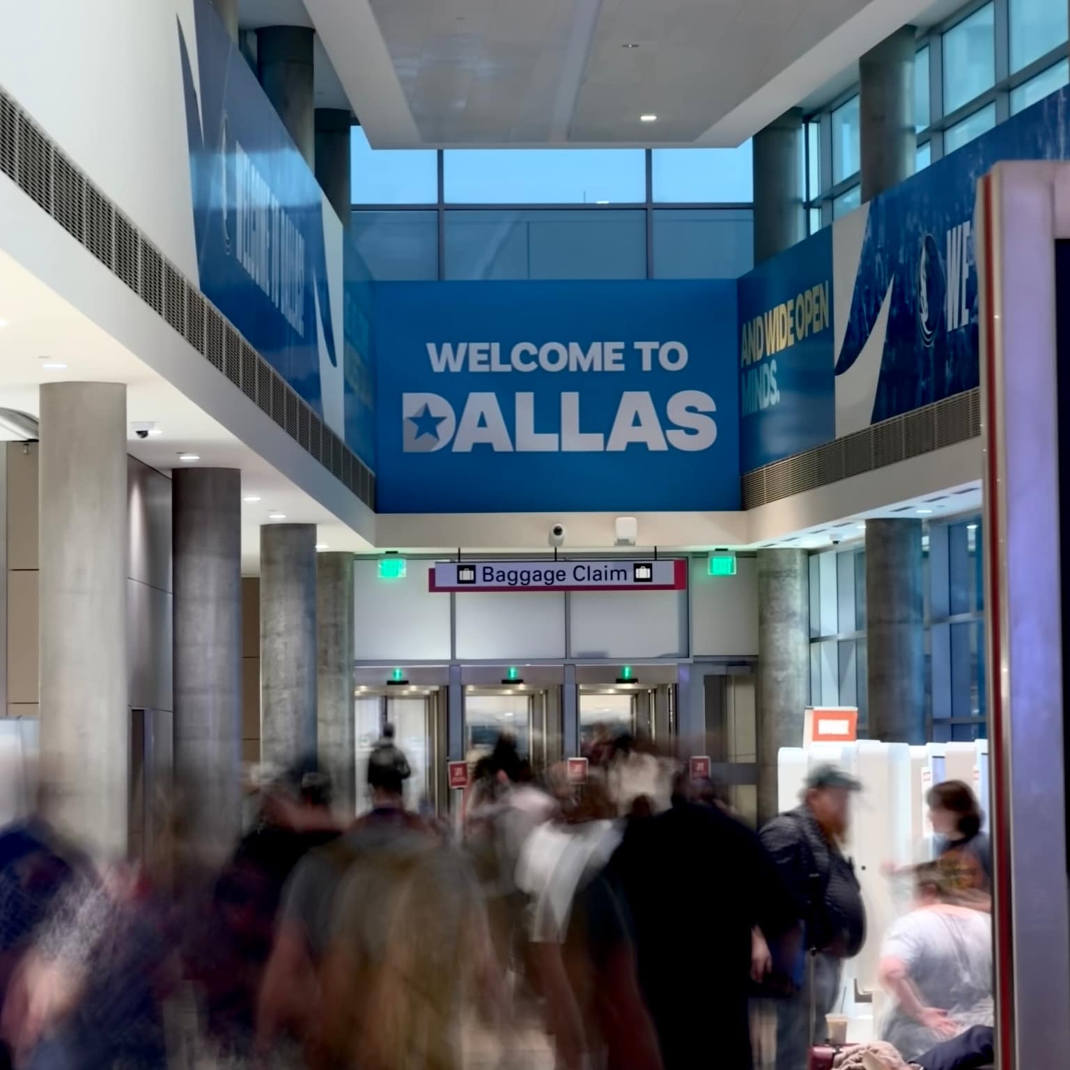 Busy airport arrivals corridor with motion-blurred travelers; large blue wall reading Welcome to Dallas and And Wide Open Minds, red Baggage Claim sign and exit lighting ahead