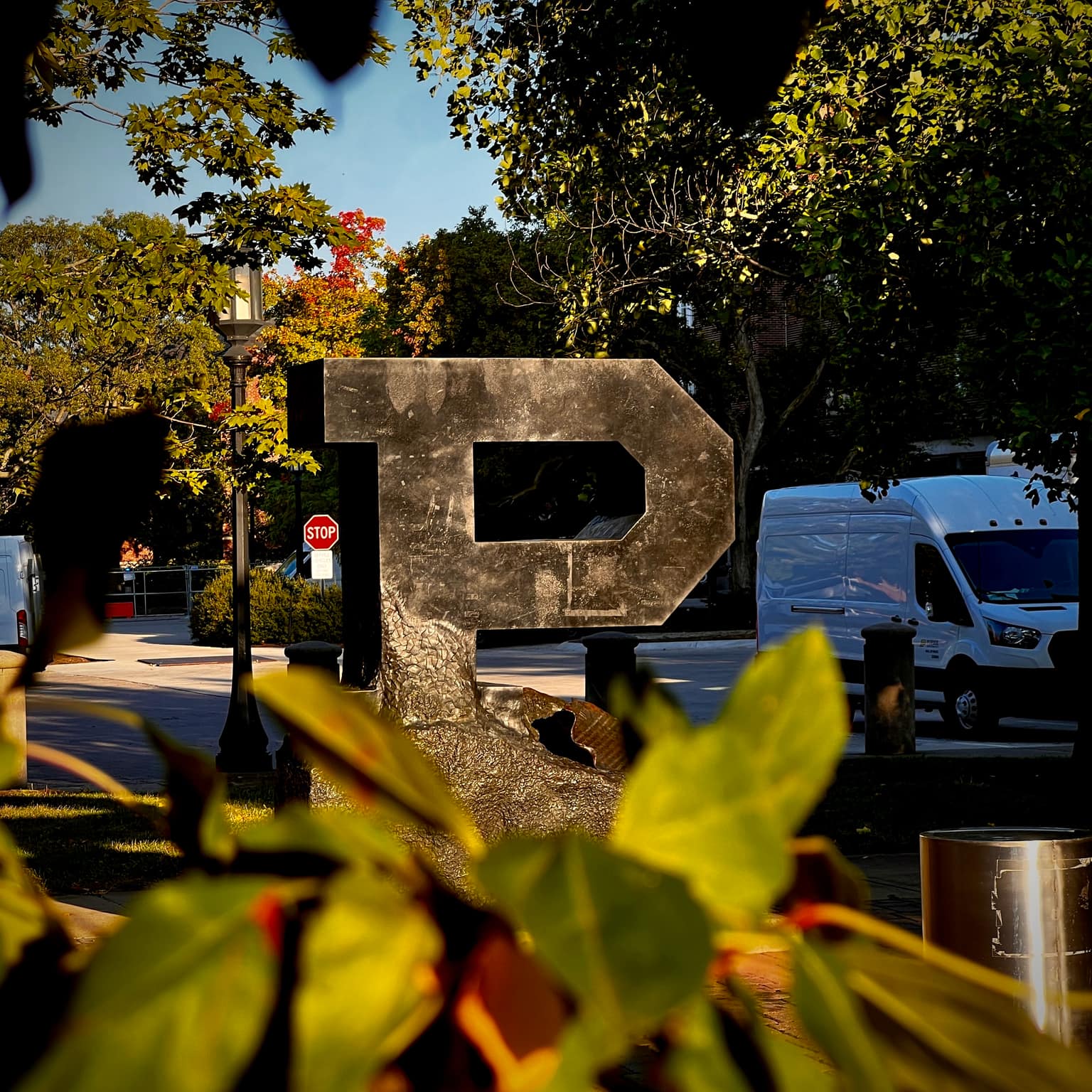 Weathered dark bronze Unfinished P campus sculpture framed by soft green leaves in the foreground, white van and lamppost beyond under sunlit trees and blue sky