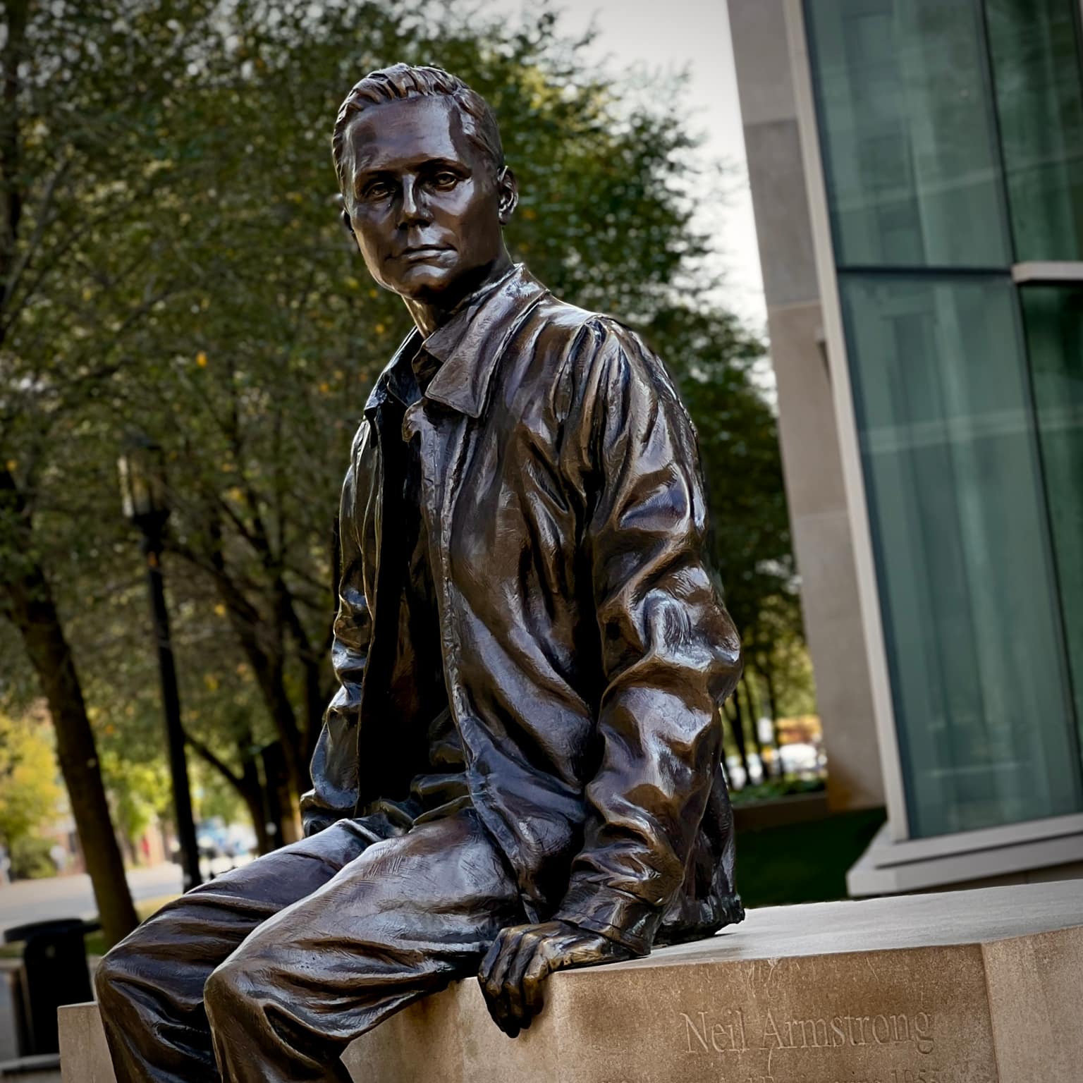 Bronze statue of young Neil Armstrong seated on a stone ledge with his name carved in the plinth, flight jacket and thoughtful pose, modern glass building and green trees behind