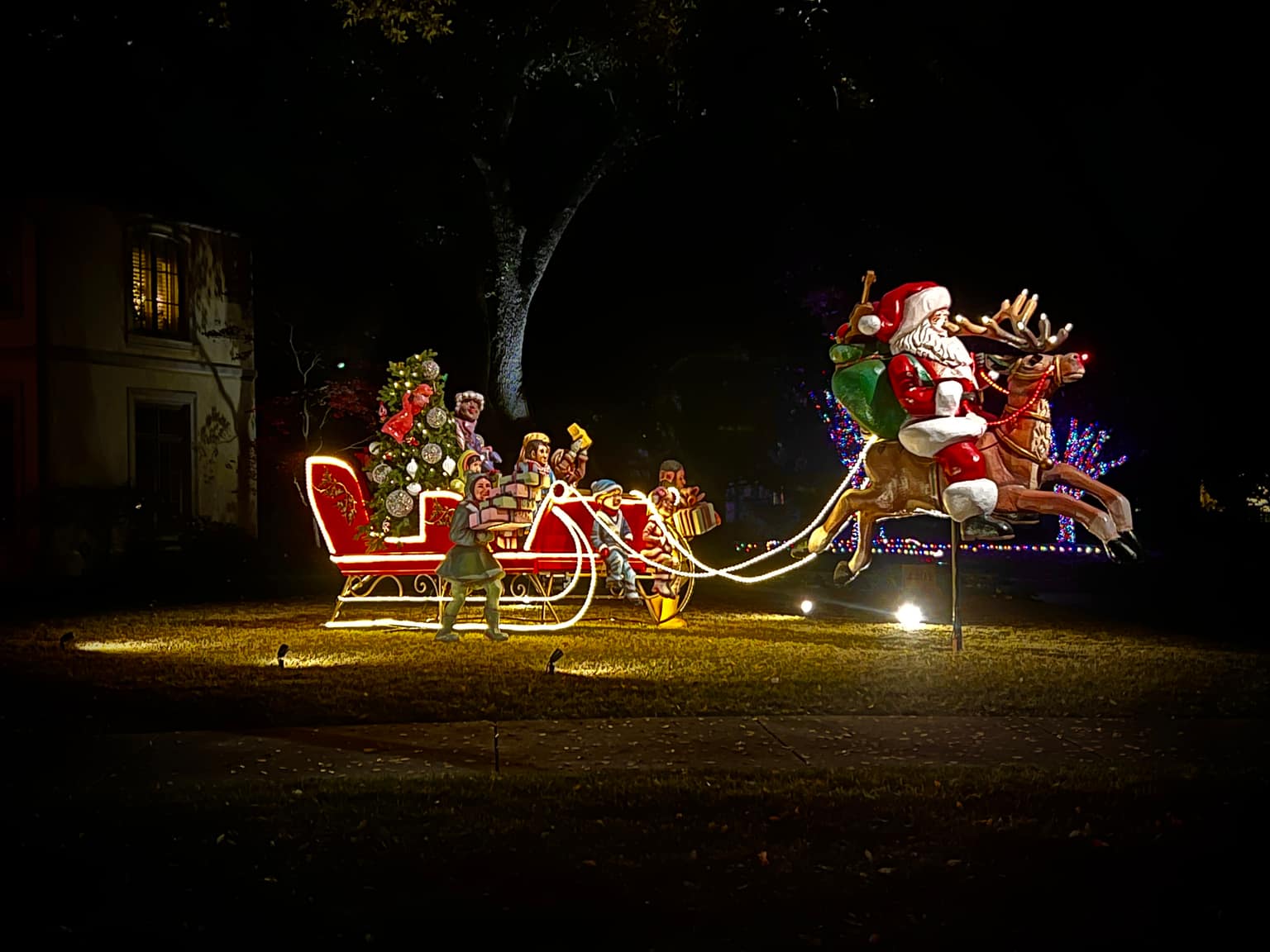 Nighttime residential lawn display: Santa on a reindeer pulls a red sleigh outlined in warm white lights, with child figures, stacked gifts, and a small lit Christmas tree