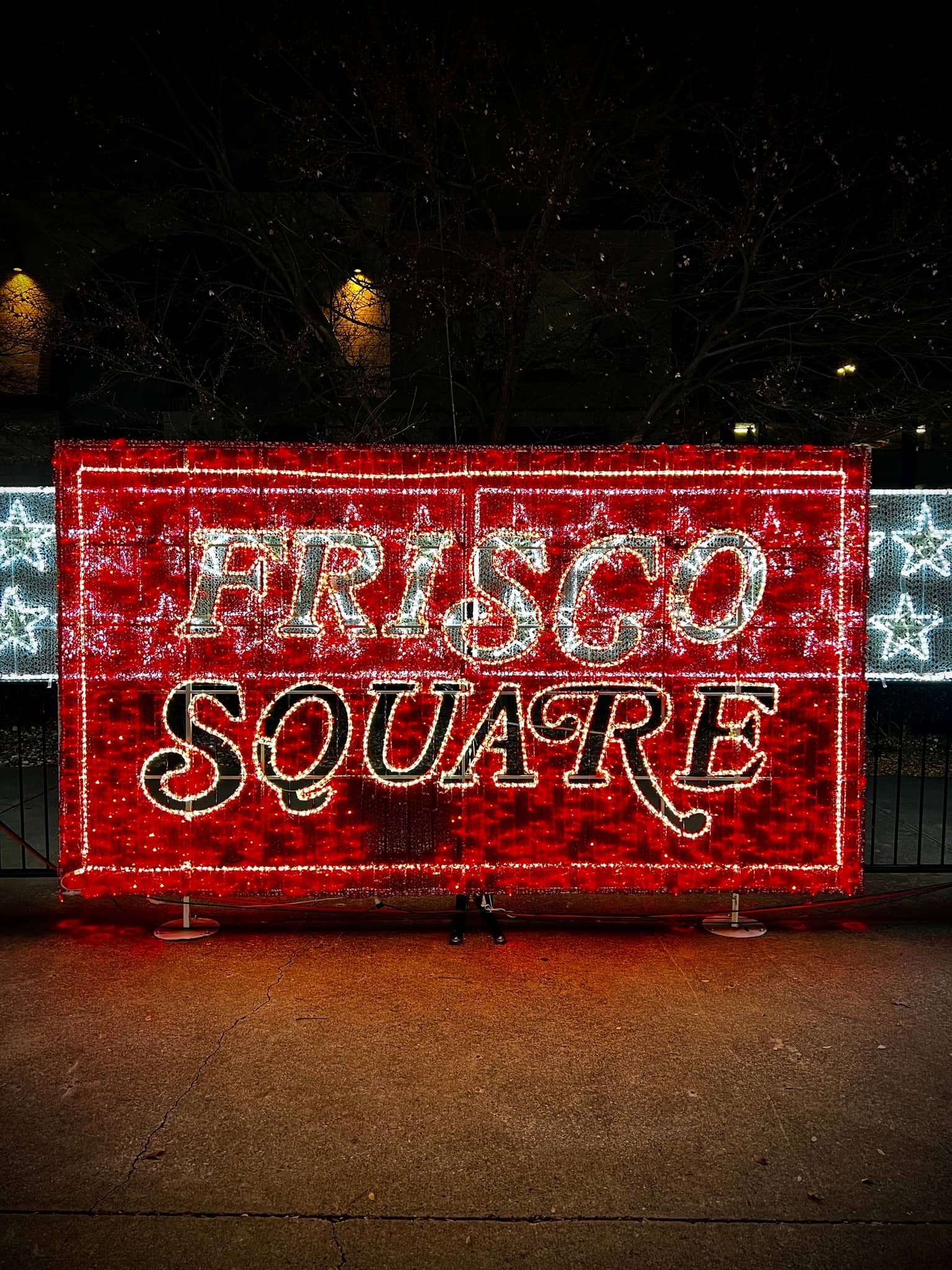 Huge rectangular holiday sign glowing red with the words Frisco Square in bright white lights, stars on either side, trees and night sky behind