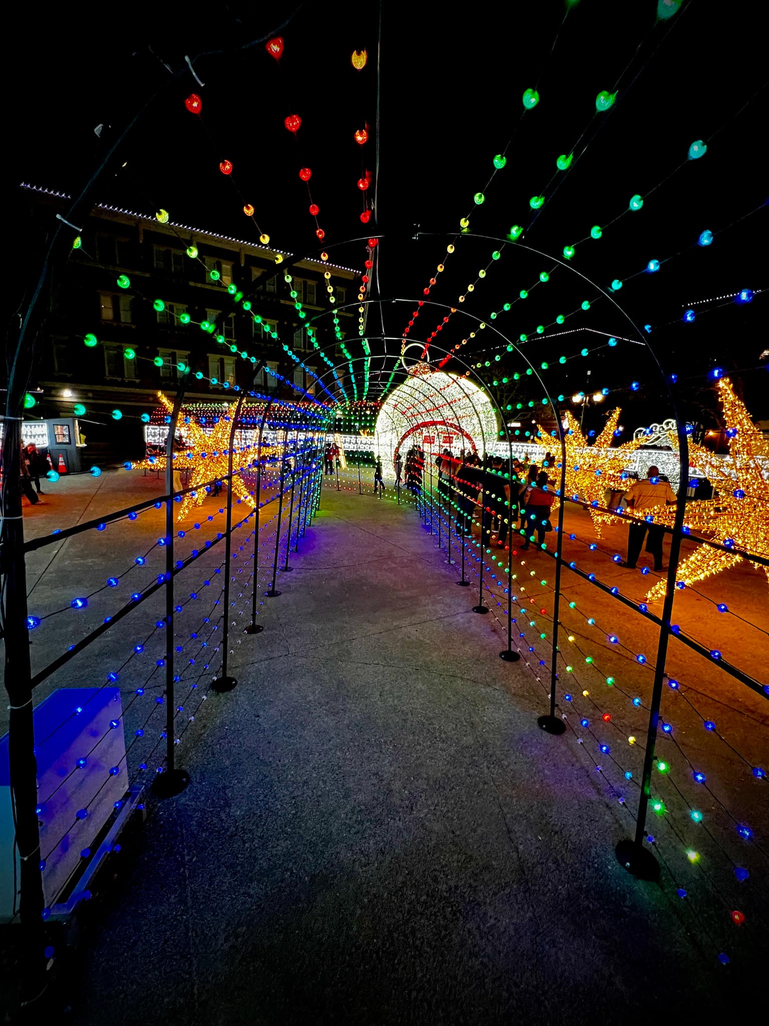 View through a long arched tunnel of rainbow-colored LED strings toward a giant walk-through sphere of white lights; large yellow star displays to the right