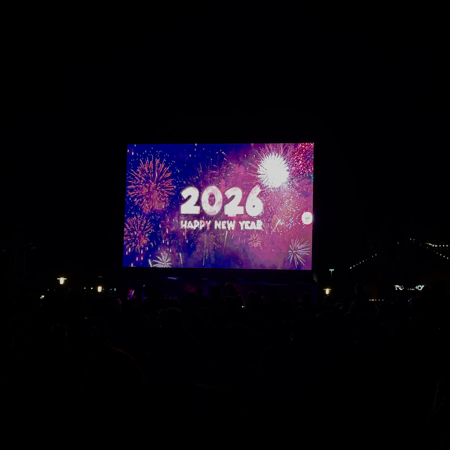 Nighttime crowd silhouettes facing a giant outdoor LED screen showing 2026 and Happy New Year with stylized fireworks