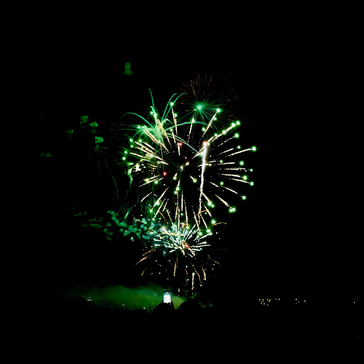 Bright lime-green fireworks bursting over a black sky while a silhouetted crowd watches; one person holds up a glowing phone to record