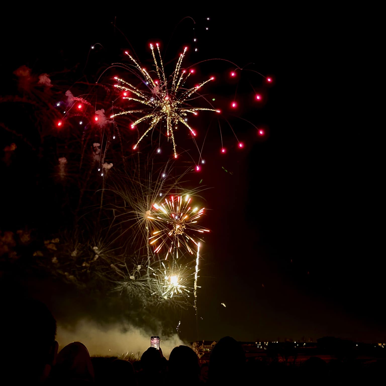 Gold and white fireworks with red spark tips above a dark crowd; a raised smartphone lights up in the foreground