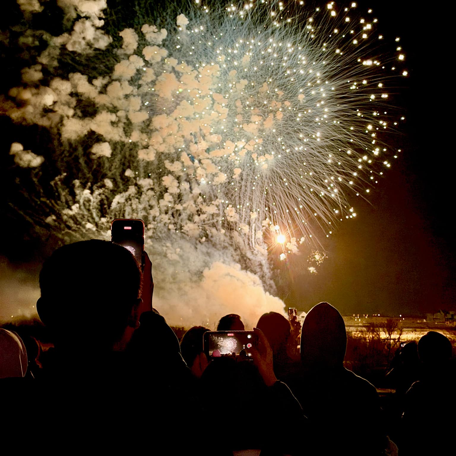 Huge white and gold firework burst and lit smoke cloud as a crowd films the show on bright phone screens