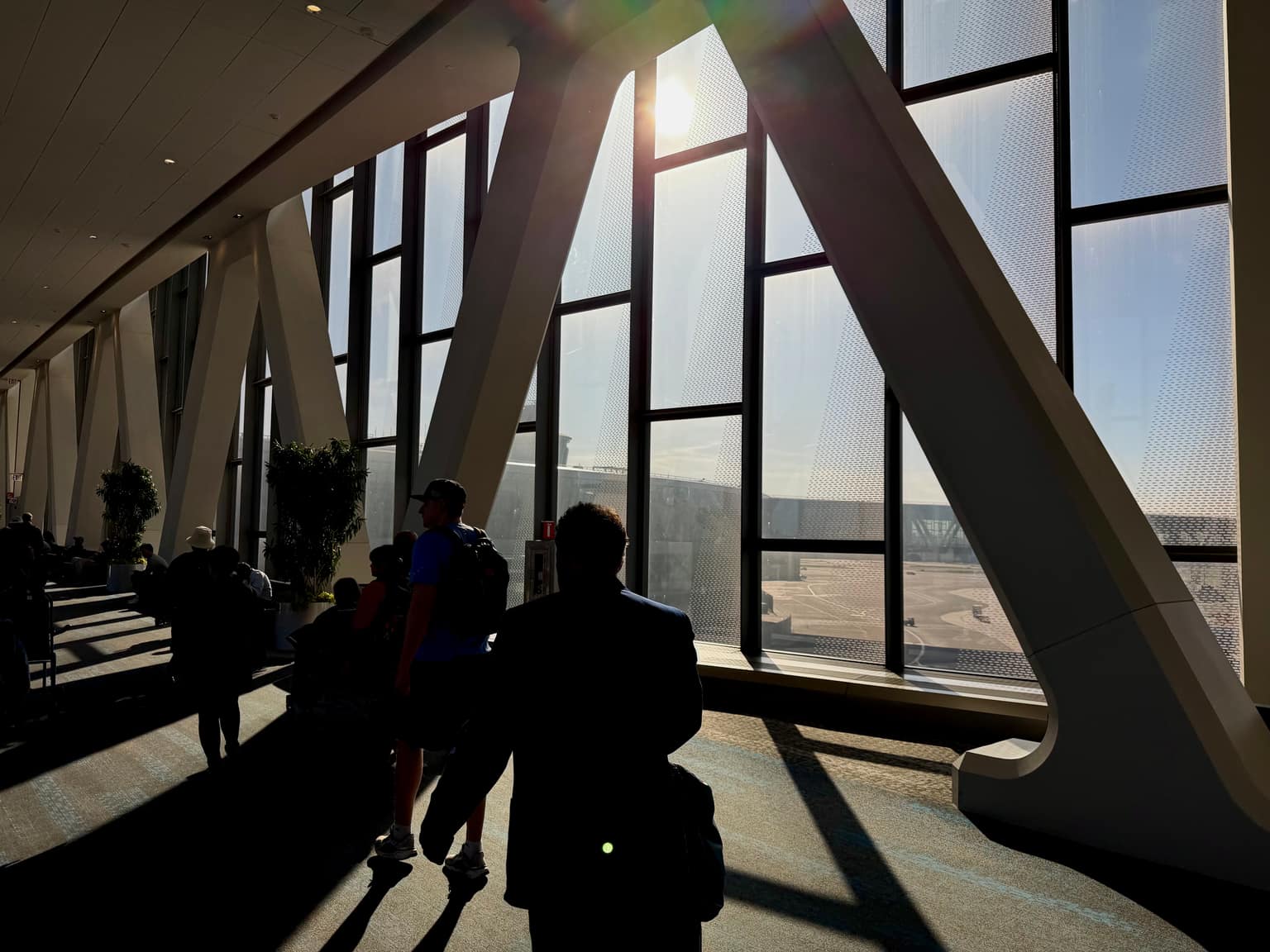 Sunlight streaming through a tall glass-walled interior with diagonal structural beams, casting long shadows across a seated crowd and the floor below