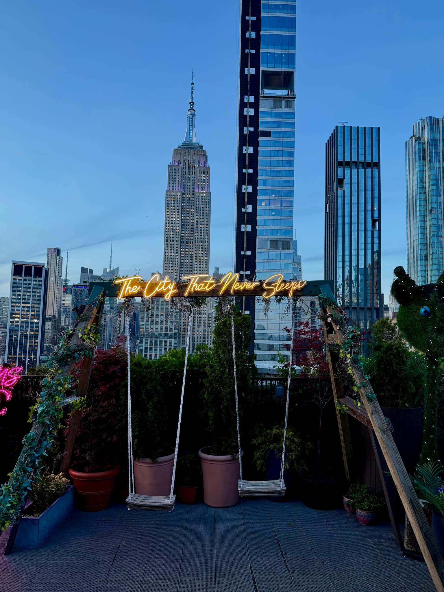 Empire State Building centered between modern towers at dusk, viewed above a rooftop deck with a glowing neon sign reading 'The City That Never Sleeps' and empty swings in the foreground