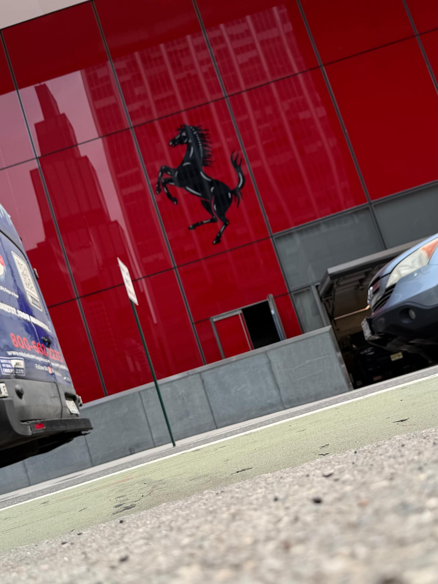 Blurred black prancing horse emblem mounted on a glossy red storefront facade, with reflections of nearby buildings and parked cars at the edges of the frame