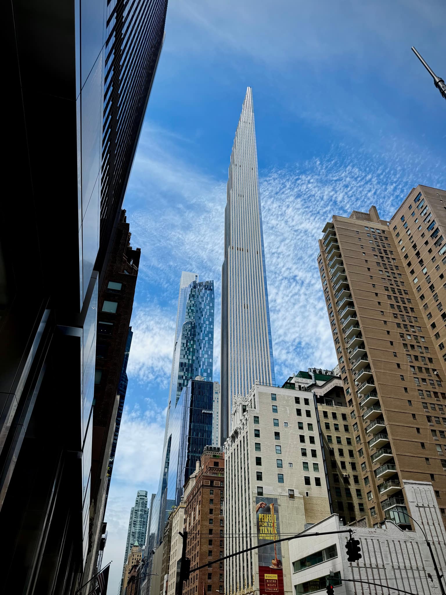 Street-level view looking up at a pencil-thin Midtown tower against a blue sky streaked with thin clouds, framed by darker glass and tan residential buildings