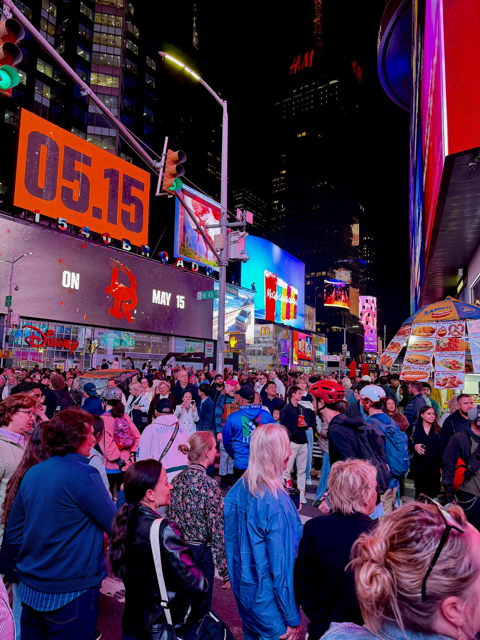 Crowded Times Square at night beneath towering digital billboards, neon storefront signs, and green traffic lights glowing over the packed intersection