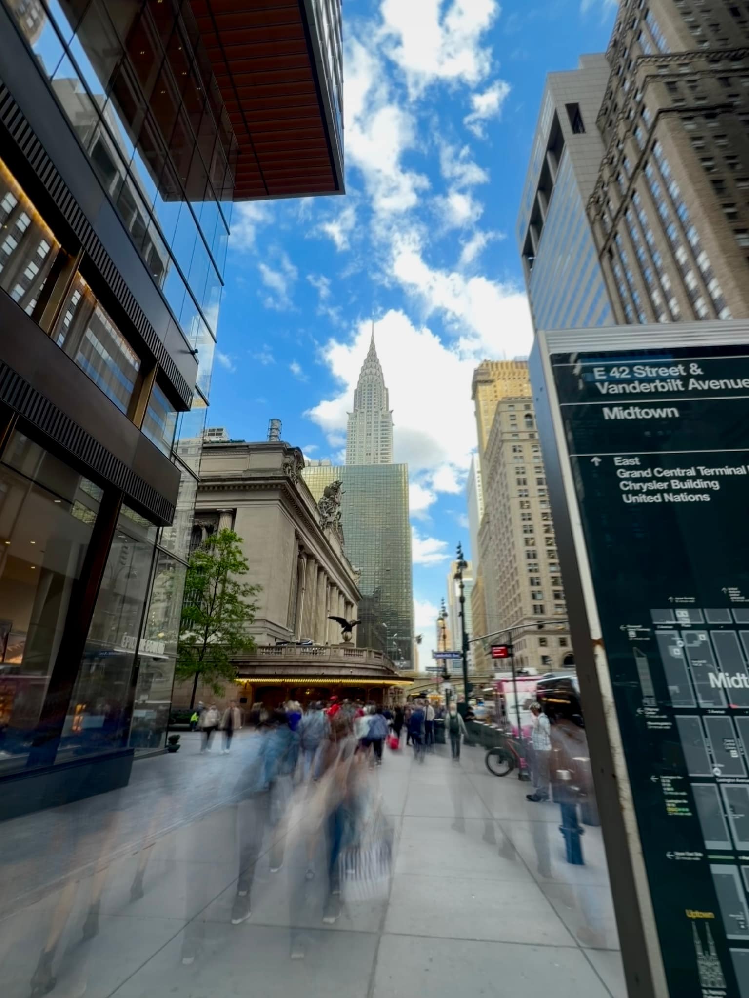 Long-exposure Midtown sidewalk scene with pedestrians blurred into motion, the Chrysler Building in the distance, and glass office facades reflecting the blue sky