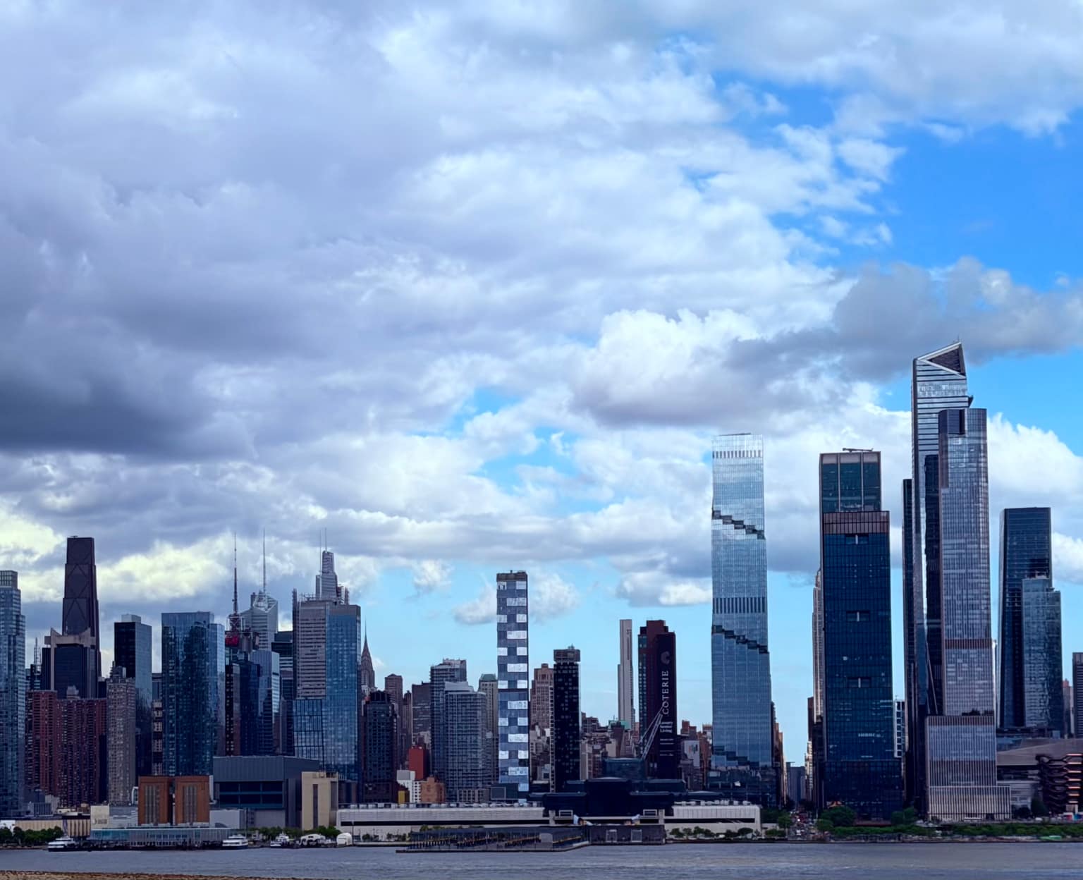 Across-the-river skyline view of Midtown Manhattan under dramatic layered clouds, with glassy high-rises stacked along the waterfront