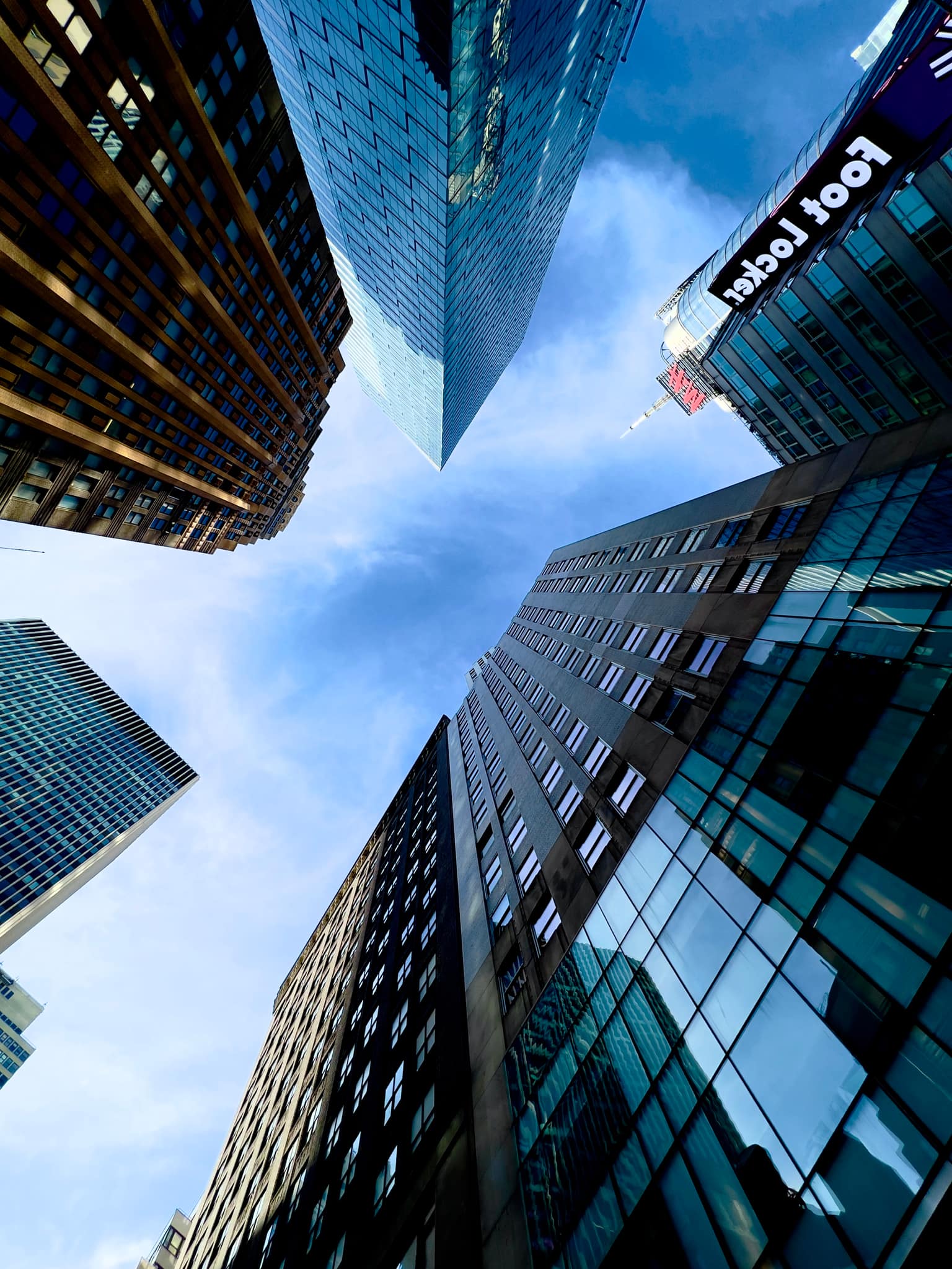 Worm's-eye view of Manhattan towers leaning inward around a patch of blue sky; glass facades and older masonry, with Foot Locker and H&M signs visible on one building