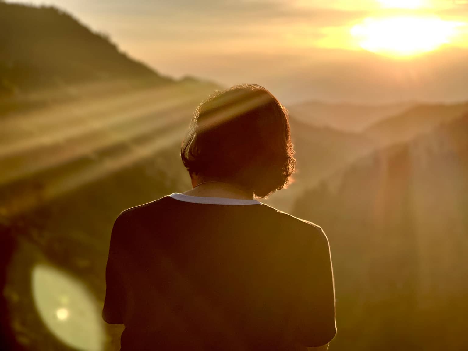 Person with dark wavy hair seen from behind, silhouetted against golden sunset light and long sunbeams over hazy blue layers of rolling mountains