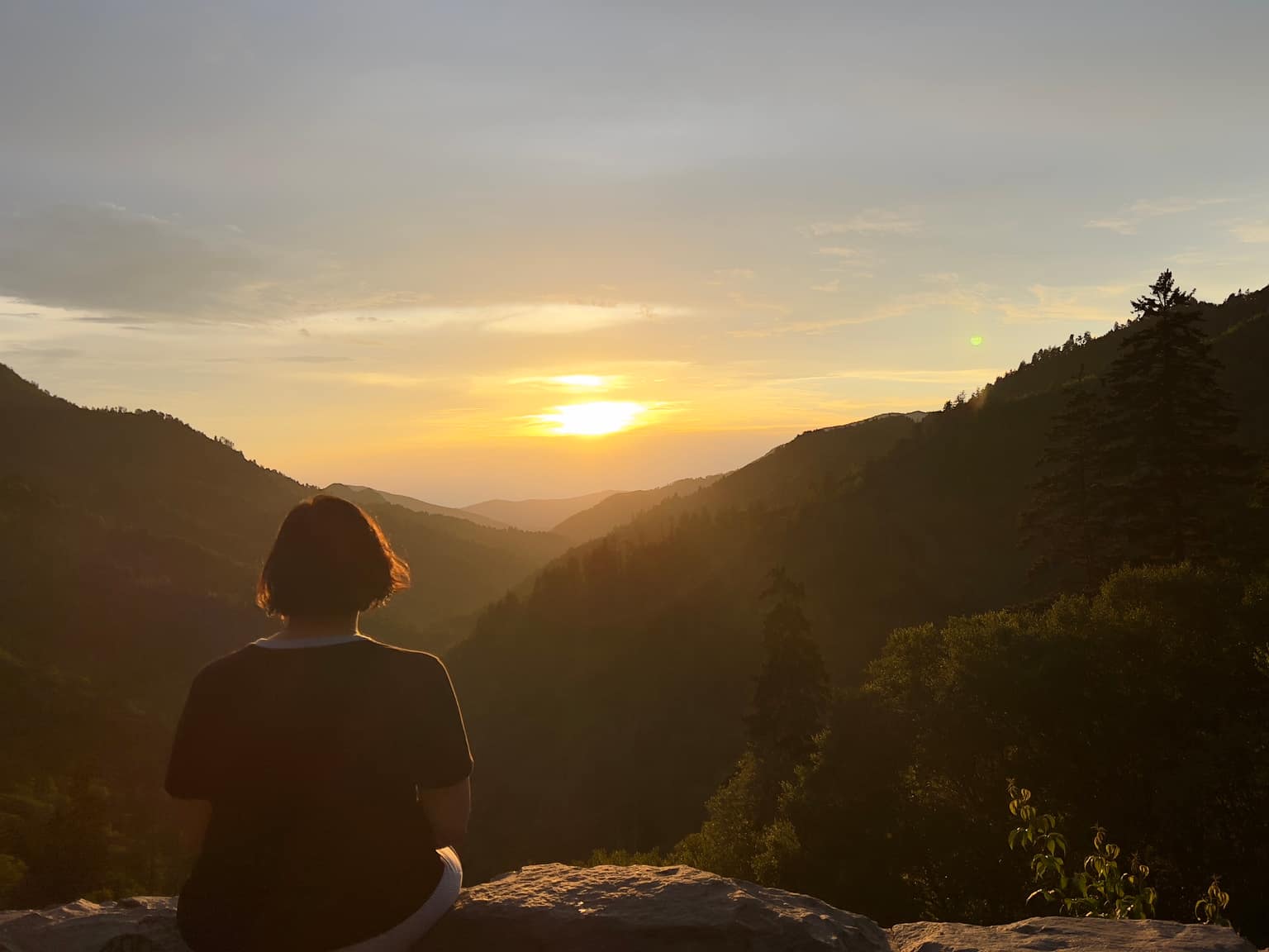 Person sitting on a rough stone wall in the foreground, facing a wide valley of forested ridges with the sun low on the horizon and warm haze in the air