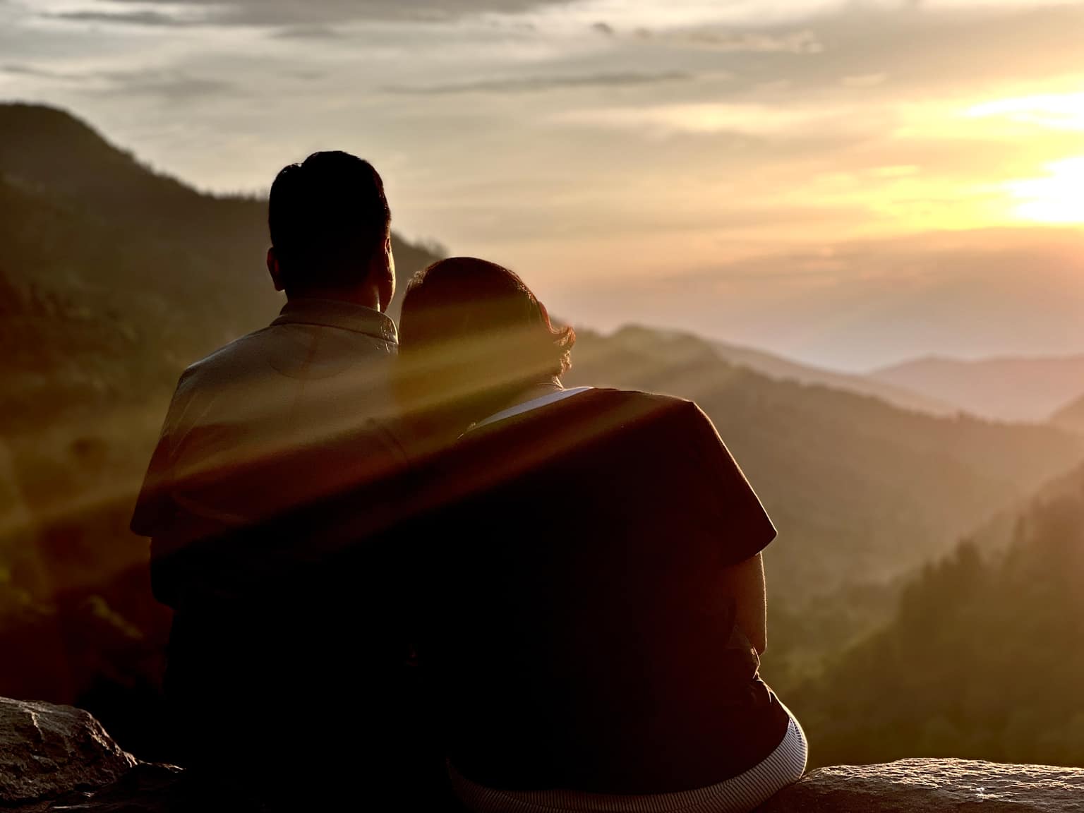 Man and woman sitting side by side on a stone ledge at sunset, her head leaning toward his shoulder, silhouetted against bright sky and receding blue mountain layers