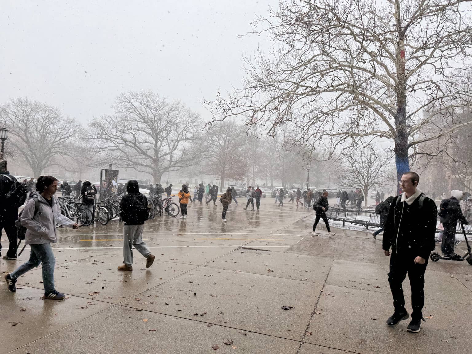 Busy paved plaza during falling snow: students in winter coats crossing slushy stone, bike racks and bare trees, grey sky and reduced visibility