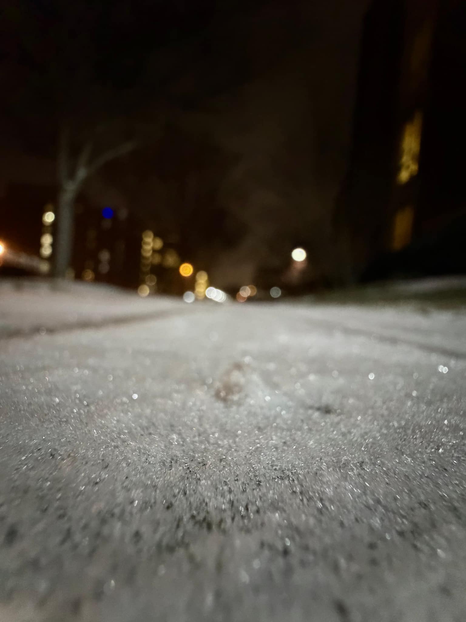 Ground-level night shot along an icy snow-covered path, sparkling granular frost in focus while bare trees and warm streetlight bokeh blur ahead