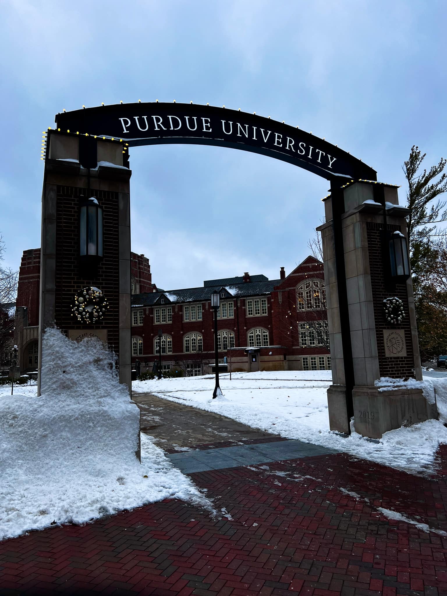Purdue University brick gateway arch with white PURDUE UNIVERSITY lettering, holiday wreaths and string lights on the span, snow on walks and piled at the pillars, red brick halls beyond