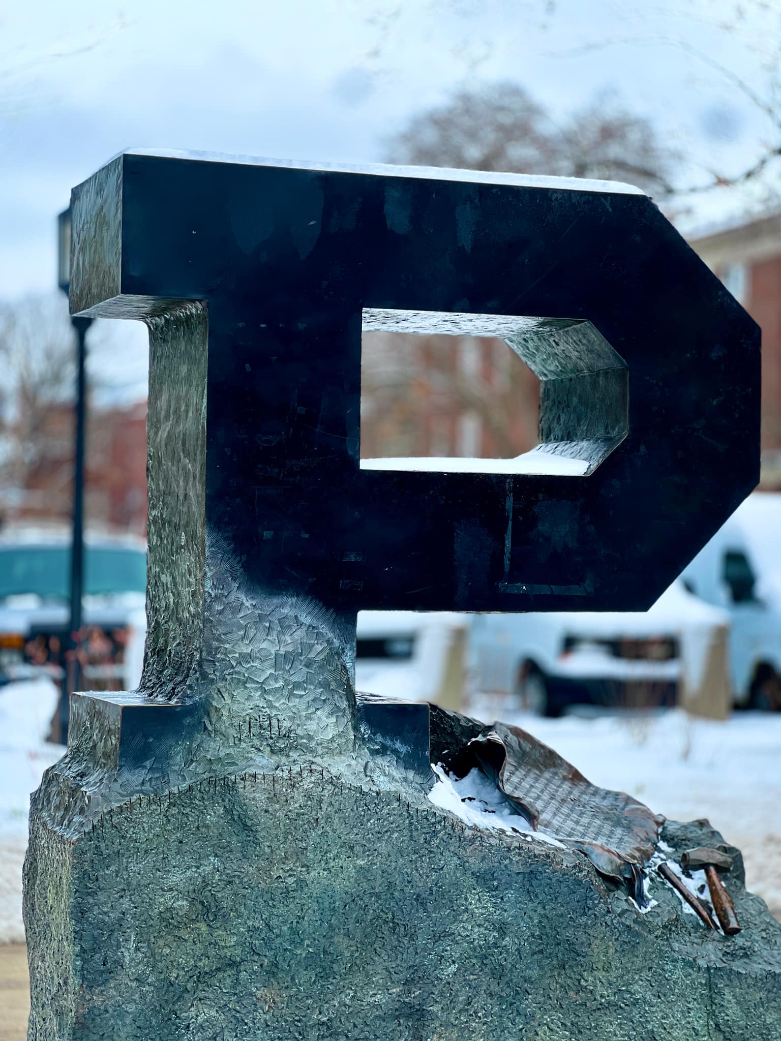 Medium shot of the Purdue Unfinished P sculpture with smooth dark upper stroke and rough stone base, bronze hammer and chisel on the plinth, light snow on surfaces and soft-focus snowy campus behind