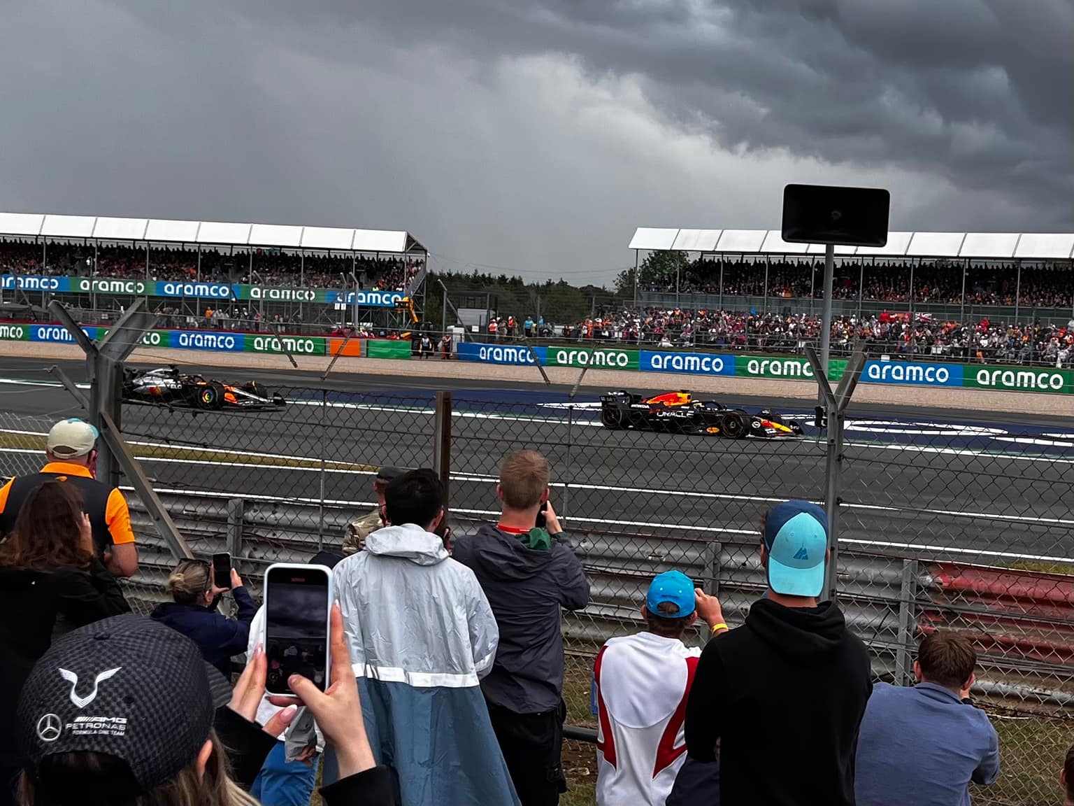 Spectators behind a fence hold phones toward the track as a Red Bull Racing car leads a papaya McLaren under storm-gray clouds; Aramco barriers and packed grandstands behind