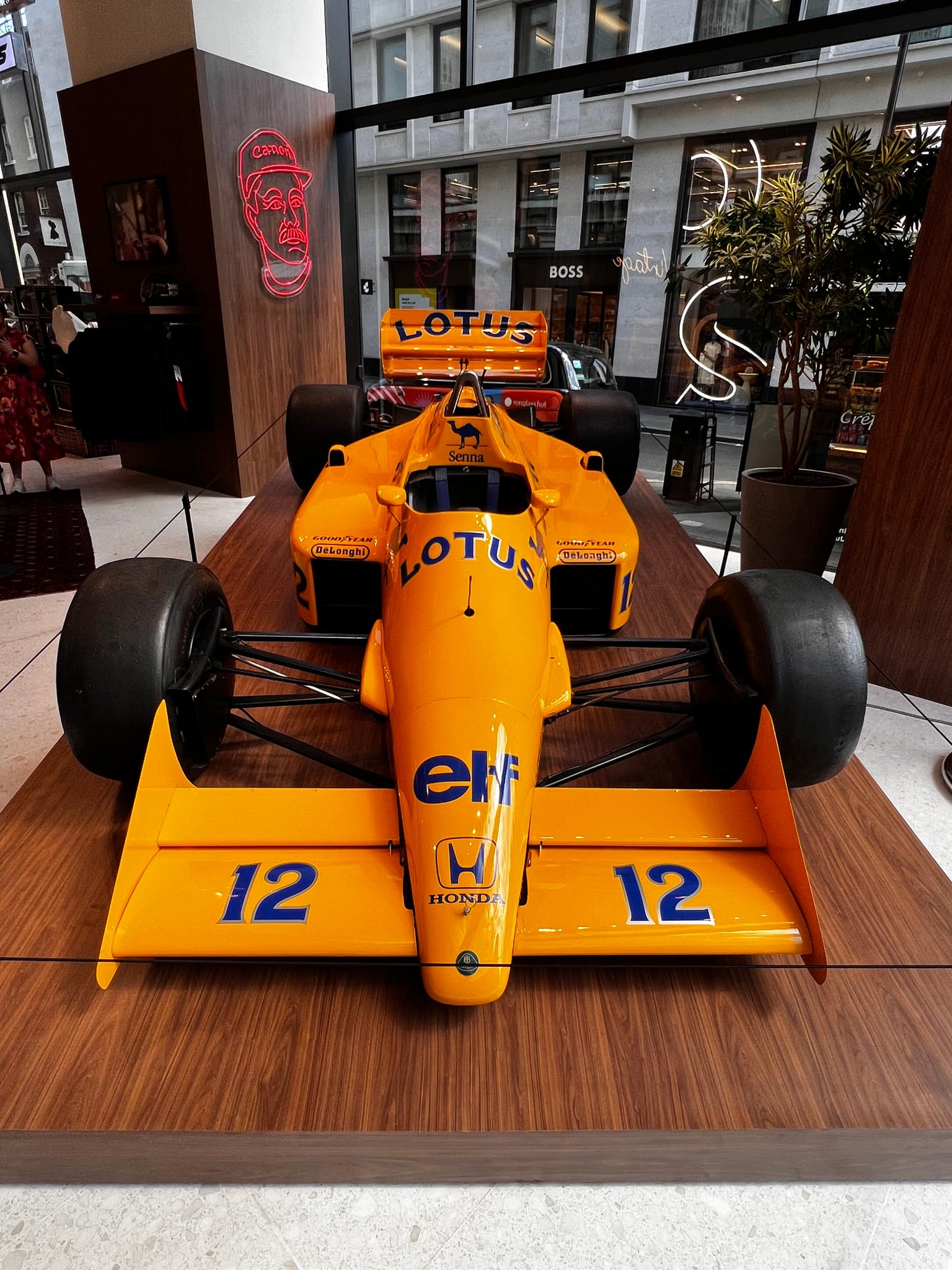 Front view of the yellow Camel-liveried Lotus 99T Formula 1 show car, number 12, Senna-era nose sponsors and Honda branding, on a wood platform inside a gallery with a BOSS storefront visible through the window