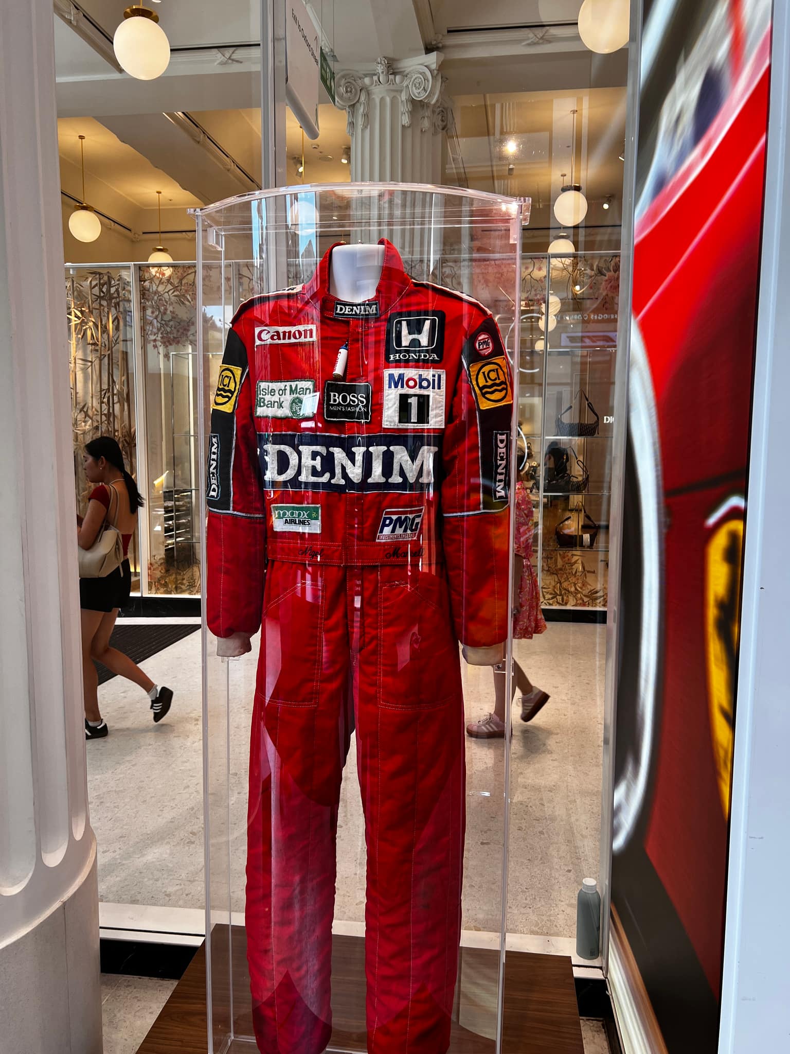 Nigel Mansell's red racing suit on a mannequin inside a tall cylindrical glass case, embroidered name on the belt and 1980s sponsor patches including Denim, Canon, Honda, Mobil 1, and Boss