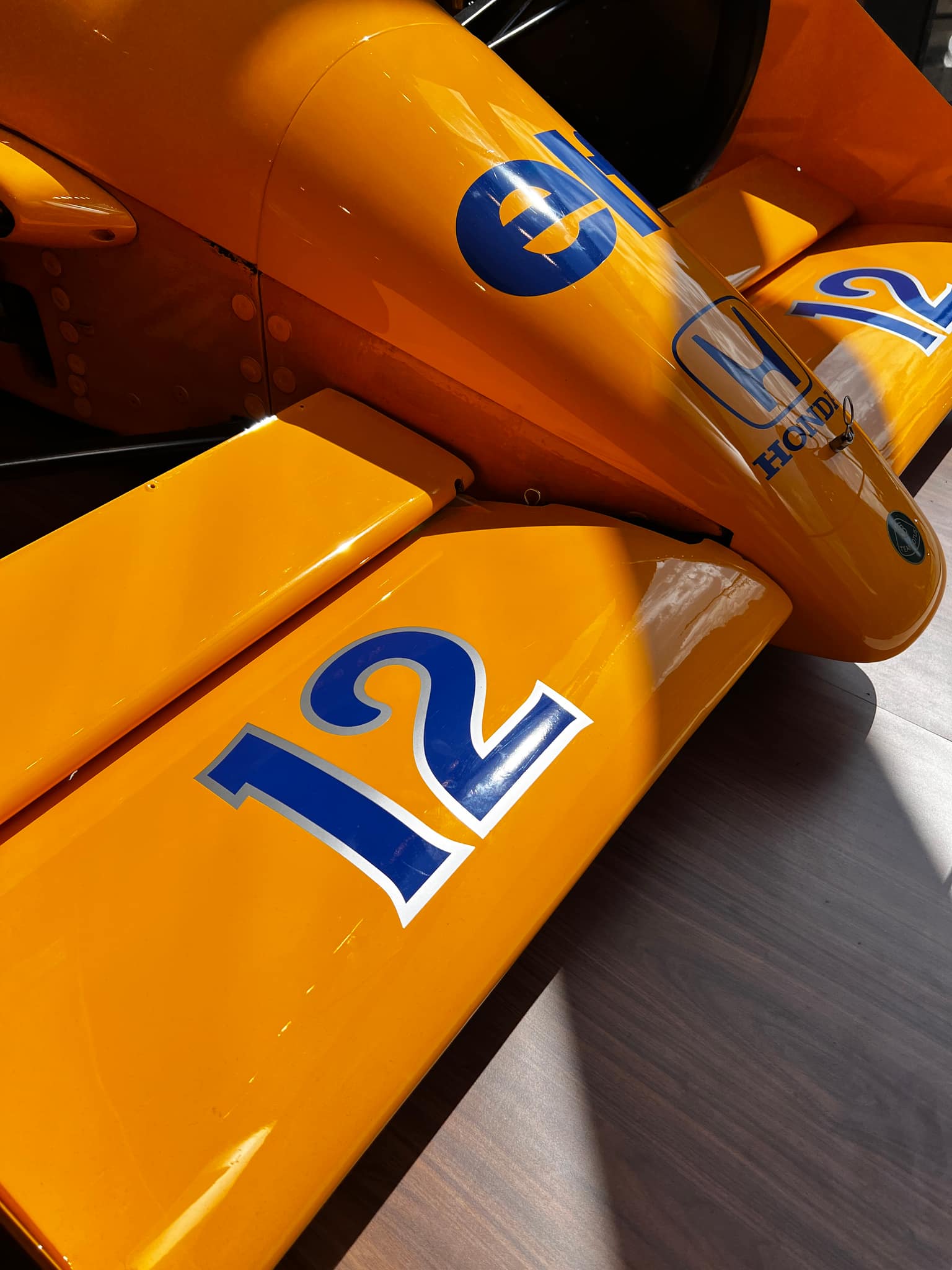 Close-up of the yellow and blue front wing and nose of a Lotus 99T, number 12, with Elf and Honda logos, glossy bodywork and rivets above dark wood flooring