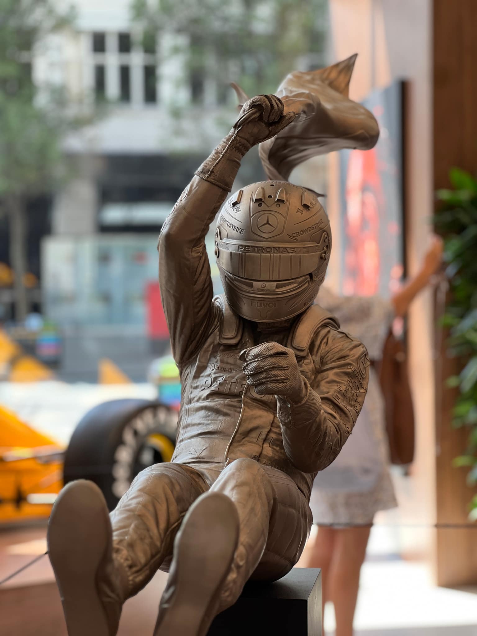 Life-sized silver statue of a Formula 1 driver in race suit and detailed helmet with Mercedes star and sponsor marks, one arm raised clutching cloth like a flag; yellow race car blur to the left