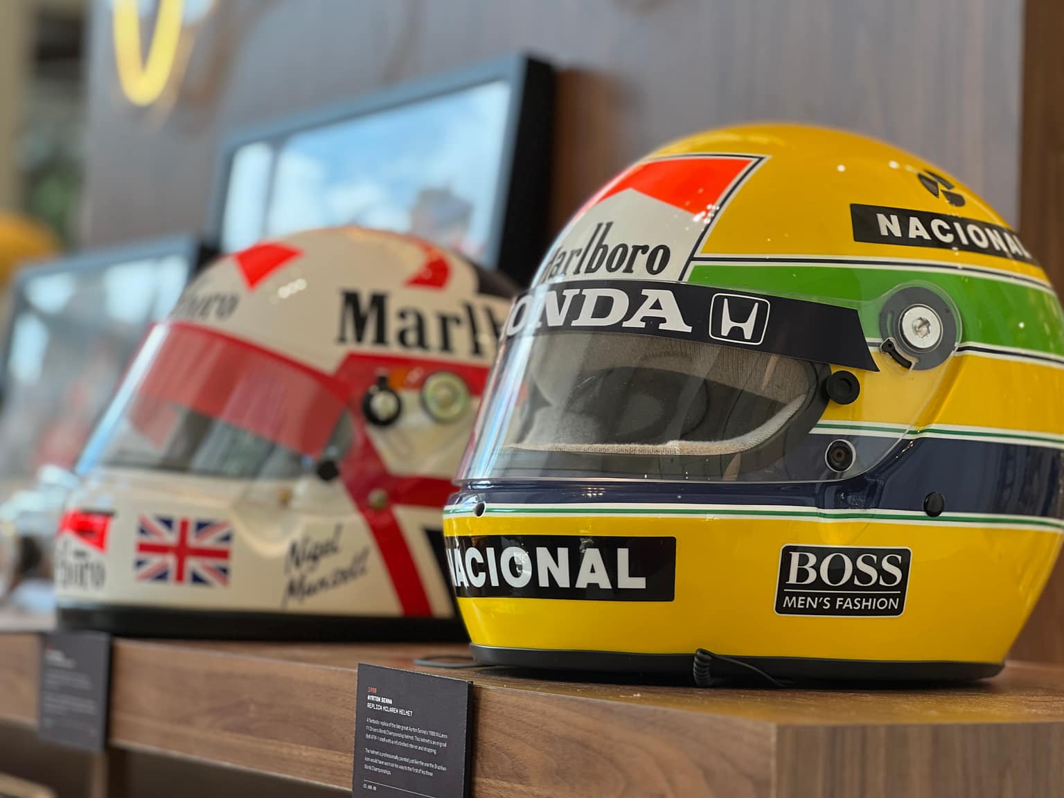 Two historic F1 helmets on a wood shelf: yellow-green-blue Ayrton Senna replica in focus with visor and sponsor decals, white-and-red Nigel Mansell helmet softly blurred behind with Union Jack detail