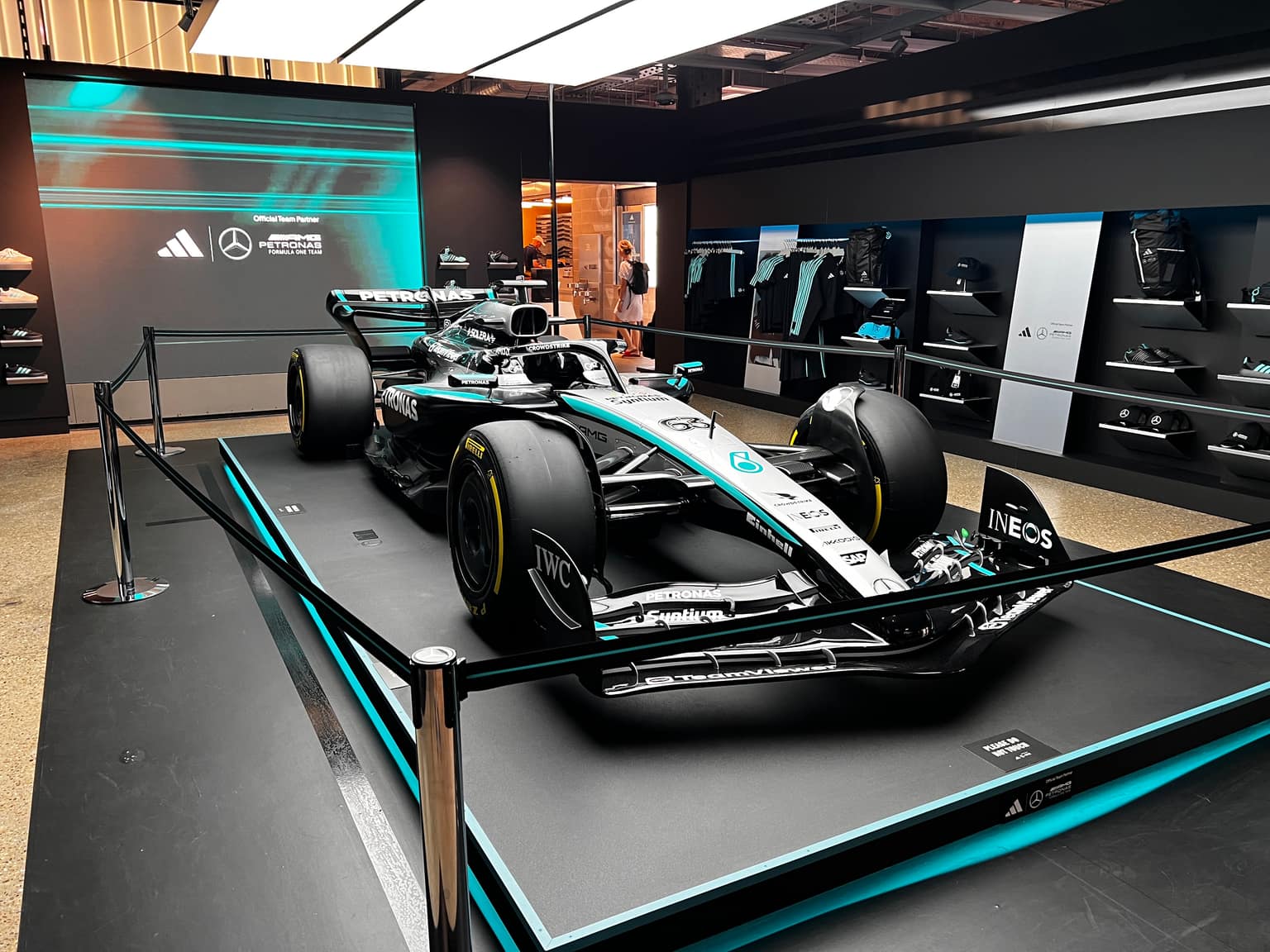 George Russell Mercedes-AMG Petronas Formula 1 car number 63 on a black raised platform with teal accent lighting; large screen with Adidas and Mercedes logos and team merchandise racks in a bright store