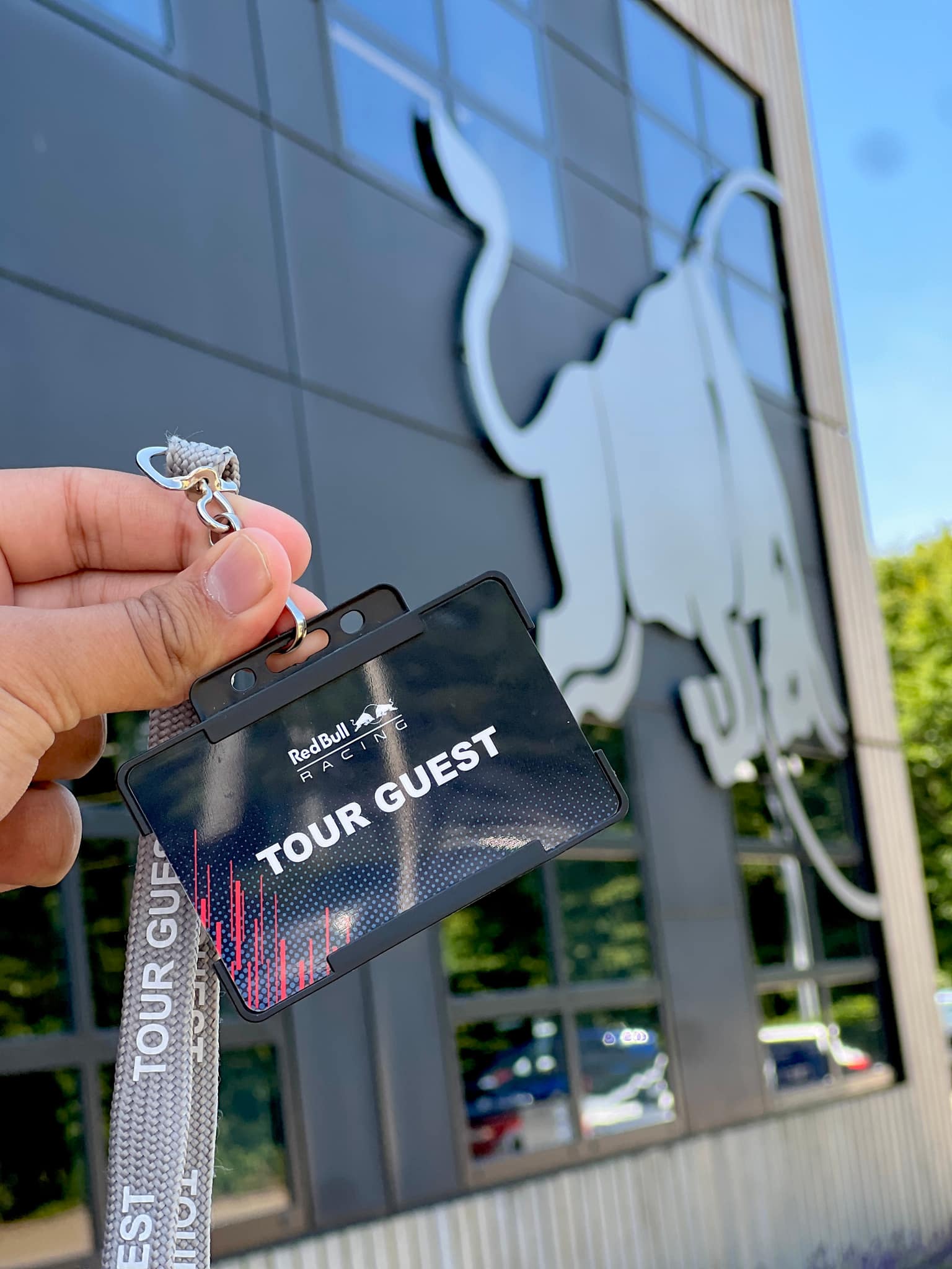 Hand holding a Red Bull Racing Tour Guest laminate on a grey woven lanyard, massive silver bull logo on the glass building facade behind under clear blue sky