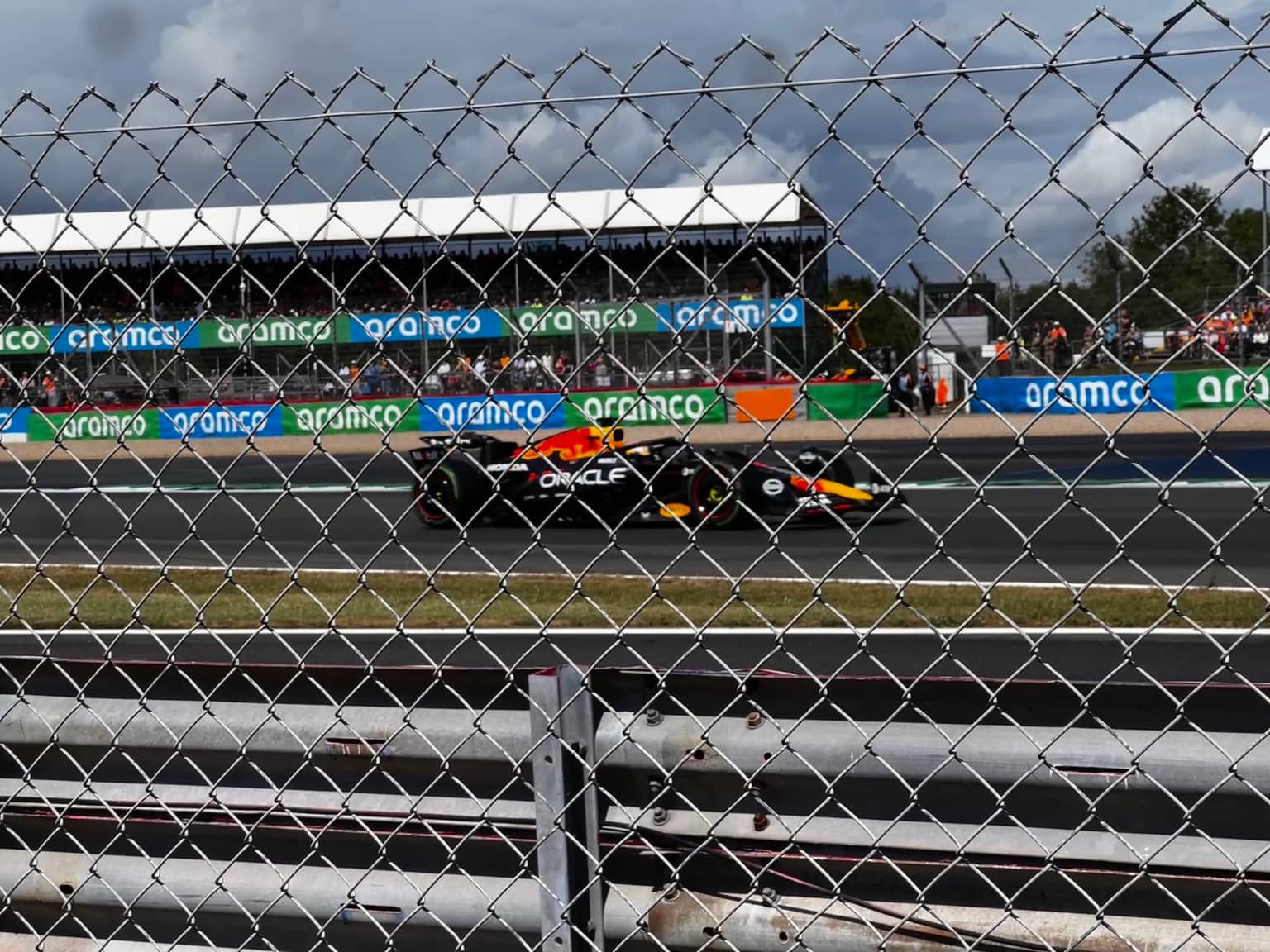 Oracle Red Bull Racing F1 car on track in profile, seen through chain-link fence and metal guardrail; green Aramco track branding and a crowded white-roof grandstand under overcast sky