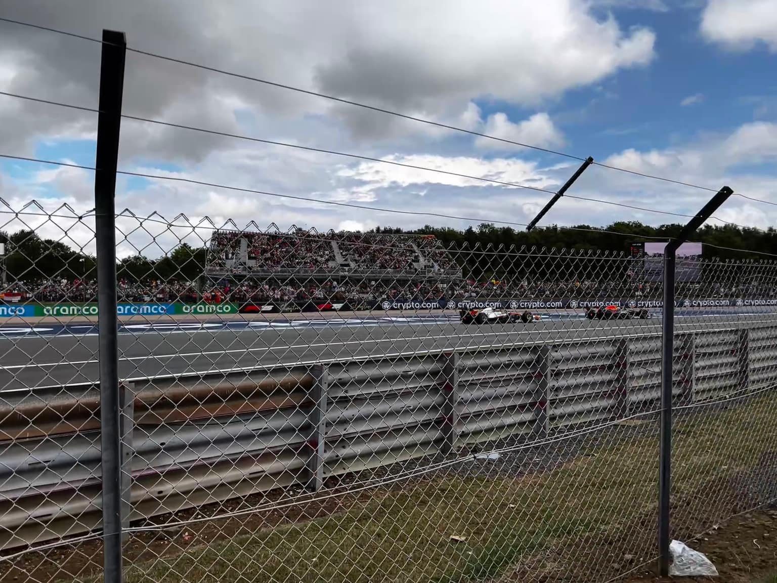 Two Formula 1 cars speed past on curbing, viewed through fence and Armco barrier; Crypto.com and Aramco banners along the wall and a full grandstand under layered gray clouds