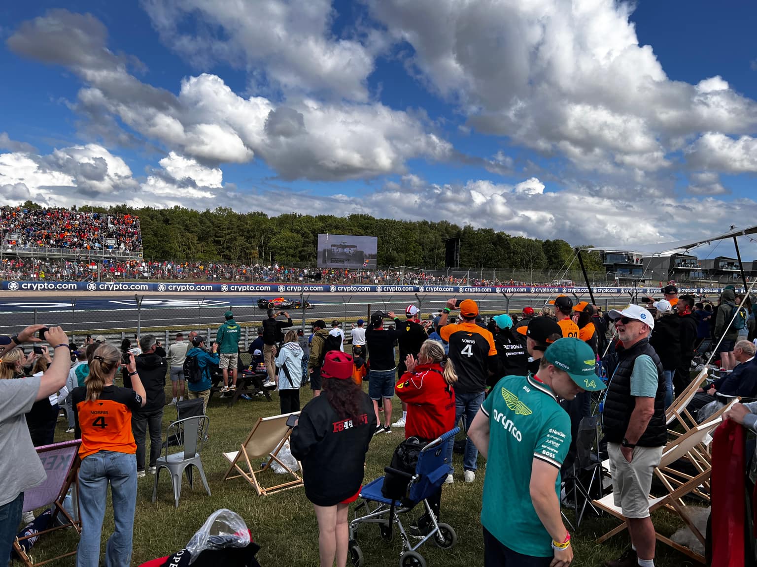 Crowd on a grassy bank in team colors—McLaren orange, Ferrari red, Aston green—as a Formula 1 car streaks along the track; crypto.com barriers, distant grandstand, and big cumulus sky